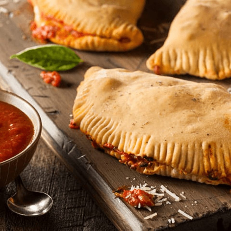 Three golden-brown Flicks calzones with tomato sauce and basil on a rustic wooden board.